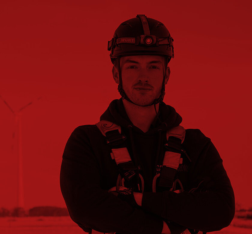Eine Person in Schutzkleidung steht mit verschränkten Armen vor drei Windkraftanlagen in einer rötlich schimmernden Landschaft.