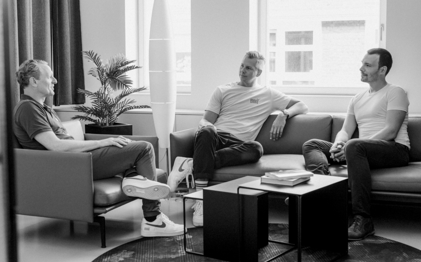 Drei Männer sitzen in einem modernen Wohnzimmer mit einem Fenster, einer Zimmerpflanze und Büchern auf dem Tisch und unterhalten sich. Die Szene wirkt ungezwungen und entspannt.