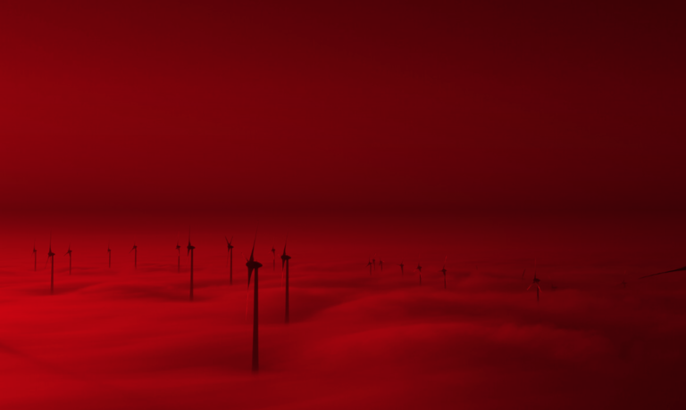 Windkraftanlagen ragen aus einer dichten Wolkendecke empor und heben sich vor einem tiefroten Himmel ab, wodurch eine surreale und futuristische Landschaft entsteht.