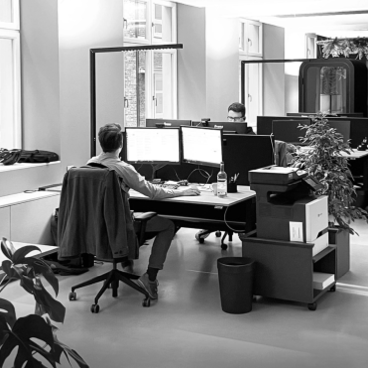 Zwei Personen arbeiten an Schreibtischen mit mehreren Monitoren in einem modernen Großraumbüro. Zu sehen sind Zimmerpflanzen und Jacken, die über den Stühlen hängen. Der Raum ist hell, da große Fenster Tageslicht hereinlassen.