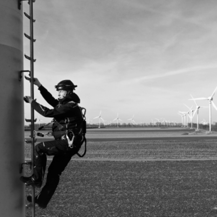 Eine Person in Schutzausrüstung klettert eine Leiter an einer Windkraftanlage hinauf; im Hintergrund sind mehrere Windkraftanlagen in einer flachen, offenen Landschaft zu sehen.